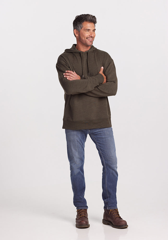 A man is standing against a plain background, wearing a Chase Hoodie - Dark Moss by Woolx made of odor-resistant Merino Wool, blue jeans, and brown boots. His arms are crossed, and he is smiling while looking to his left.