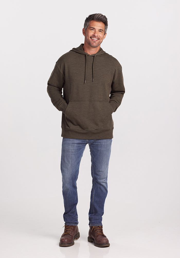 A man stands against a plain background, smiling with his hands in the front pocket of his dark moss Chase Hoodie by Woolx. He is also wearing blue jeans and brown boots, exuding the perfect blend of comfort and style for any outdoor adventure.