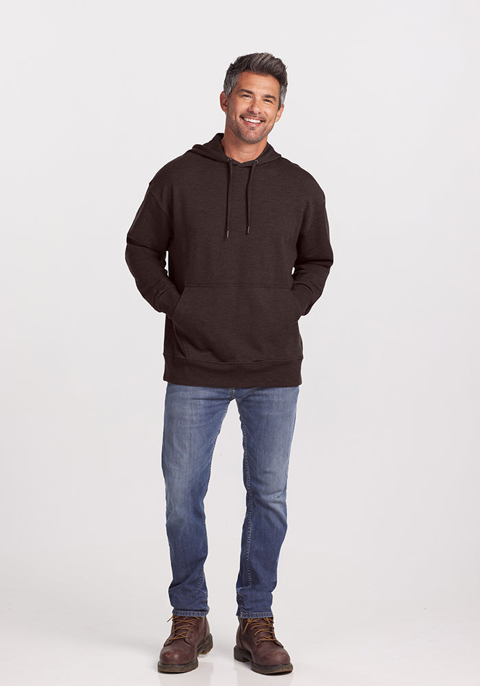 A man stands smiling against a plain background, wearing a Maple Melange Chase Hoodie from Woolx with his hands in its front pocket. He has short, gray-tinged hair and a beard. He is also wearing blue jeans and brown boots.