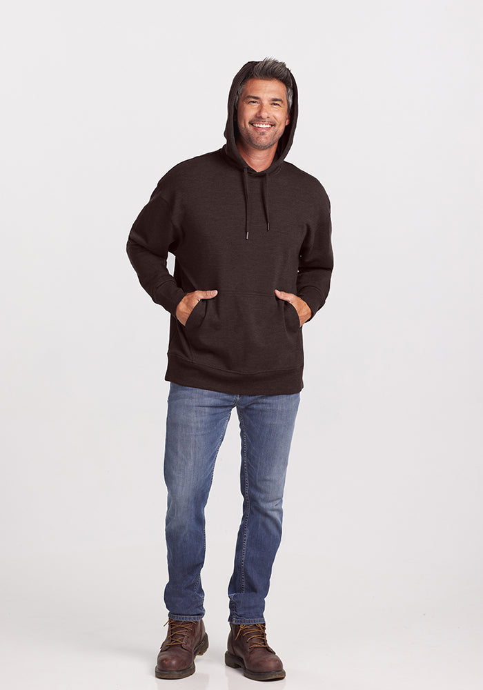 A person wearing a Maple Melange Chase Hoodie from Woolx, blue jeans, and brown boots is standing against a plain white background. They have their hands in the hoodie pockets and are smiling at the camera.