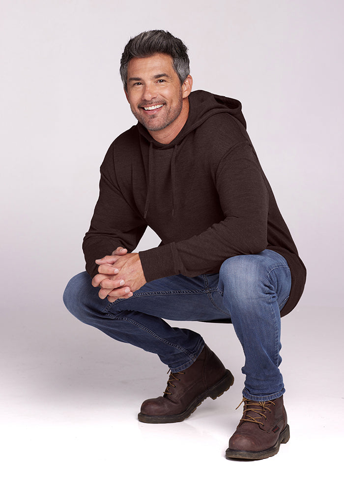 A man with short gray hair and a beard is crouching and smiling at the camera. He is wearing a dark brown Chase Hoodie - Maple Melange by Woolx, blue jeans, and brown boots. The background is plain white.