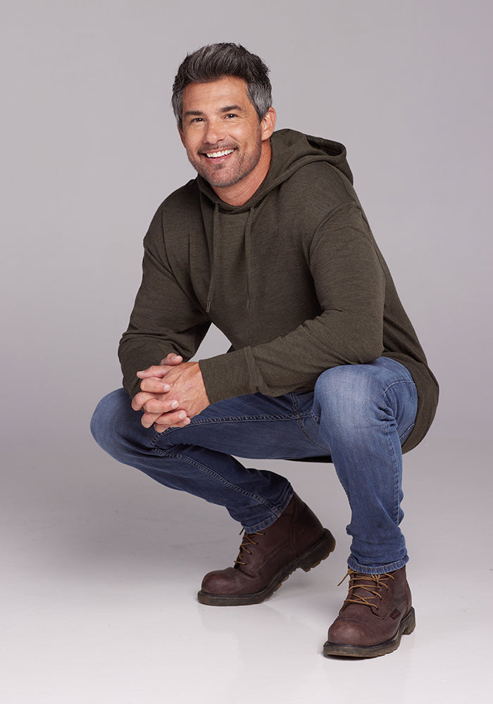 A smiling man with salt-and-pepper hair, wearing a Woolx Chase Hoodie in Dark Moss, blue jeans, and brown boots, crouches down with his hands clasped in front of him against a plain, light-gray background.