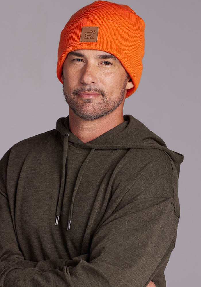 A man with a short beard and mustache is wearing a bright orange Woolx Baylor Beanie and an olive-green hoodie. He is looking directly at the camera with a slight smile. The background is a plain, neutral gray.