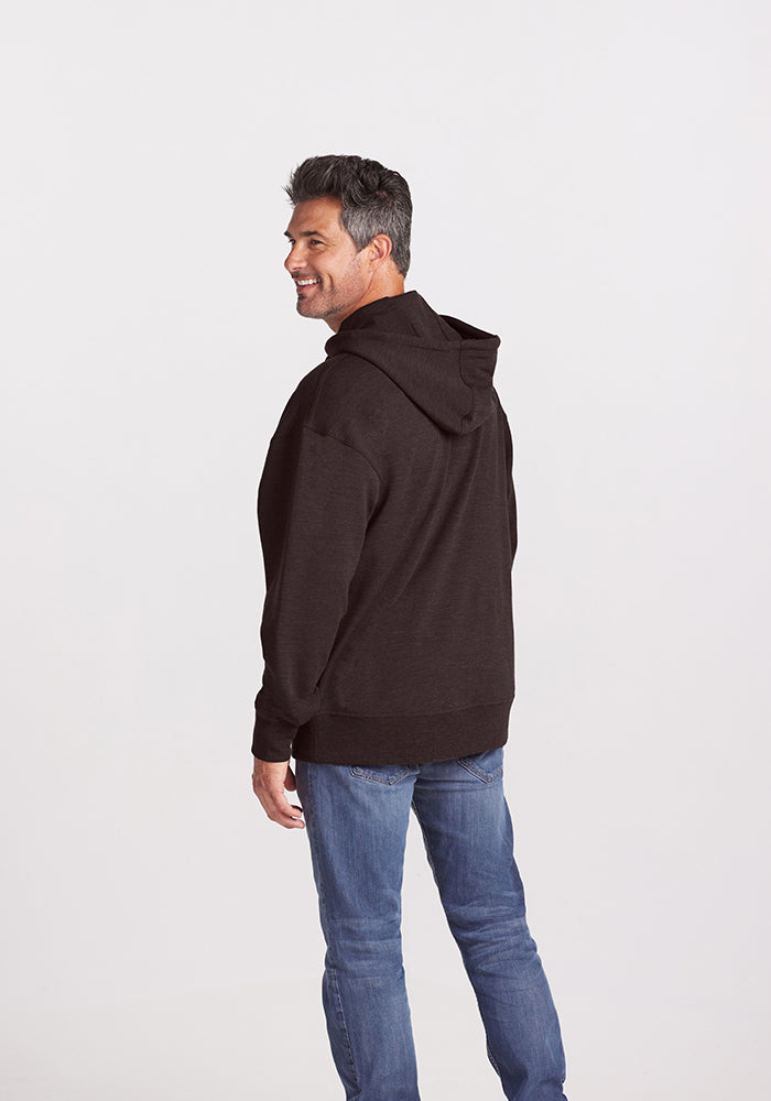 A man with short, dark hair is seen from behind, looking over his shoulder and smiling. He is wearing a dark Maple Melange Chase Hoodie by Woolx made of Merino wool and blue jeans. The background is plain and light-colored, likely a studio setting.