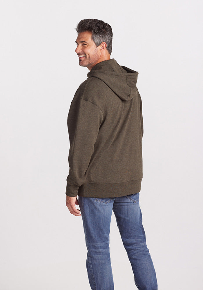 A man with short dark hair, smiling and slightly turned to the side while facing away from the camera, is wearing a Woolx Chase Hoodie in Dark Moss made from soft Merino wool and blue jeans against a plain white background.