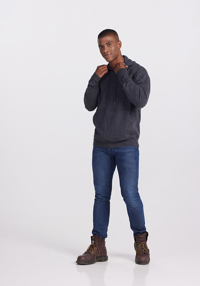 A person stands confidently against a plain background, dressed in Woolx’s Chase Hoodie in Pebble Grey Melange, paired with blue jeans and brown boots. Their hands are slightly raised as they adjust the hood strings of the warm merino wool hoodie, and they maintain a neutral expression.