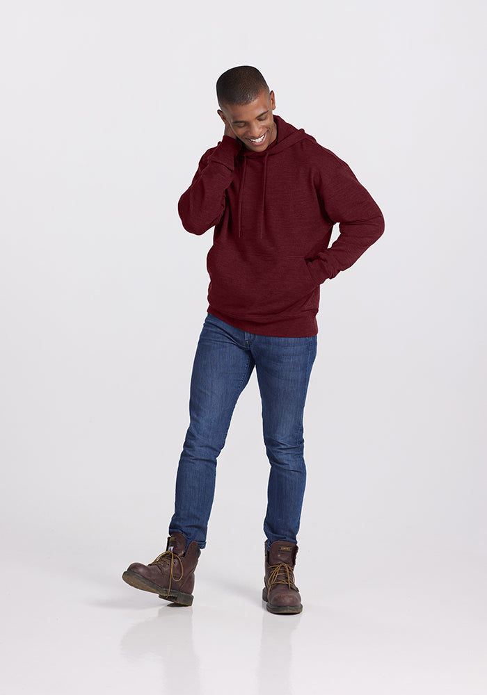 A person is standing and smiling with their head tilted slightly and one hand touching their head. They are wearing a Cranberry Melange Chase Hoodie by Woolx, blue jeans, and brown boots. The background is plain and light-colored.