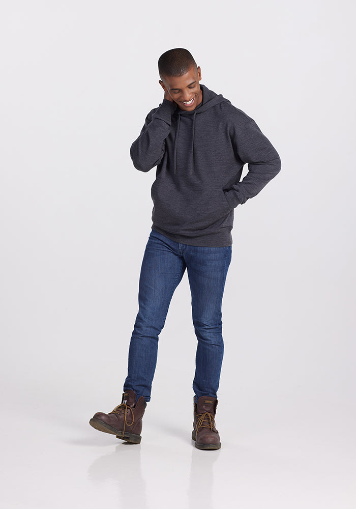 A person with short hair stands against a plain background. They are wearing a Woolx Chase Hoodie in Pebble Grey Melange, blue jeans, and brown boots. They have one hand touching the side of their head and are smiling while looking slightly downward.