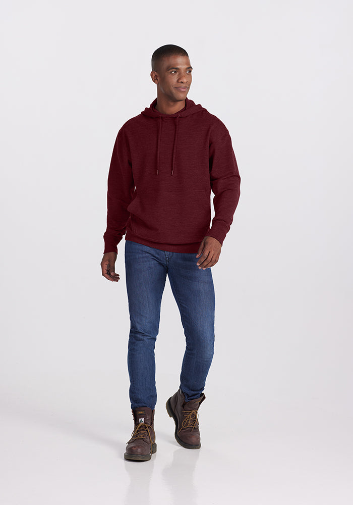 A person is standing against a plain background, wearing a Cranberry Melange Chase Hoodie from Woolx, blue jeans, and brown boots. They are looking slightly to the side with a relaxed expression and one hand in their pocket.