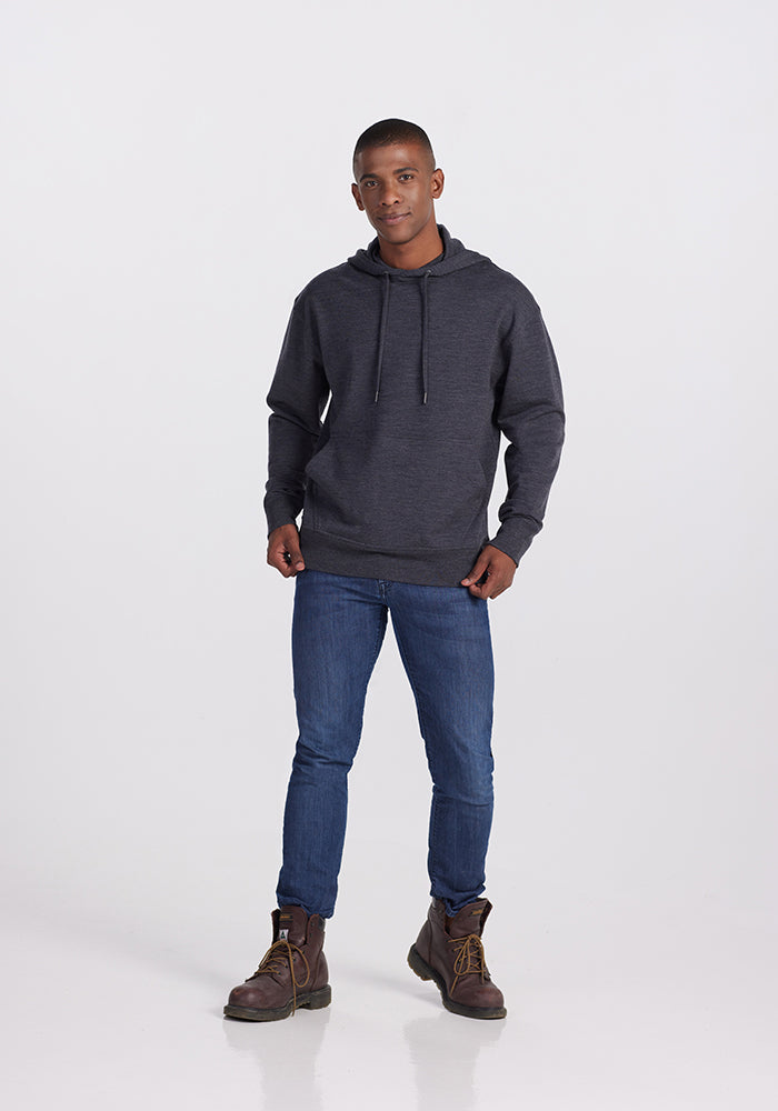 A person stands against a plain white background, wearing a Woolx Chase Hoodie in Pebble Grey Melange with drawstrings, blue jeans, and brown work boots. The person has a neutral expression and stands with legs slightly apart and hands resting on thighs.