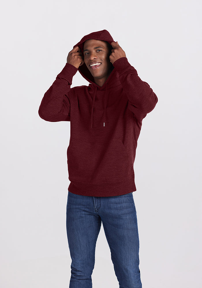 A person stands against a plain background, smiling and adjusting the hood of their Cranberry Melange Chase Hoodie from Woolx. They are wearing blue jeans and have their hands raised to hold the edges of the hood over their head.