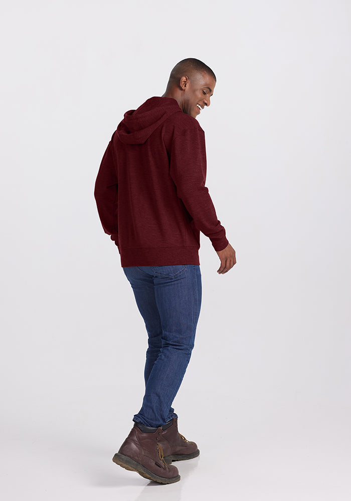 A person wearing a Woolx Chase Hoodie in Cranberry Melange, blue jeans, and brown boots is standing and smiling while looking over their shoulder. The individual is facing slightly away from the camera. The background is plain and light-colored.