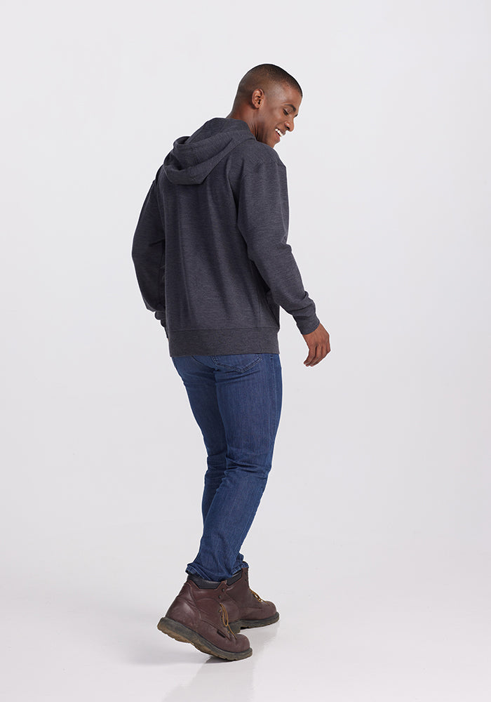 A man stands against a plain white background, facing slightly away from the camera. He is smiling with one hand raised, wearing a dark gray Woolx Chase Hoodie in Pebble Grey Melange, blue jeans, and brown ankle boots.