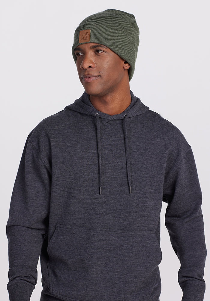 A person wearing a Woolx Chase Hoodie in Pebble Grey Melange and an olive green merino wool knitted beanie, which features a brown patch on the front, is looking slightly to the side. The background is plain and light-colored.