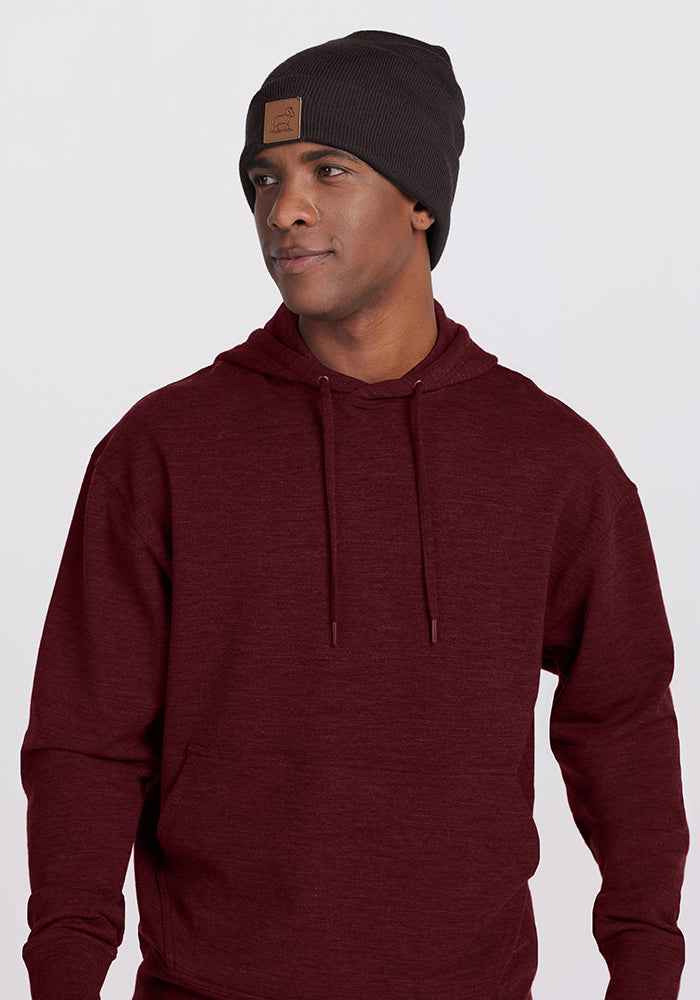 A person is wearing a Chase Hoodie - Cranberry Melange from Woolx and a dark gray beanie with a square brown patch on the front. They are looking off to the side against a plain, light-colored background. 