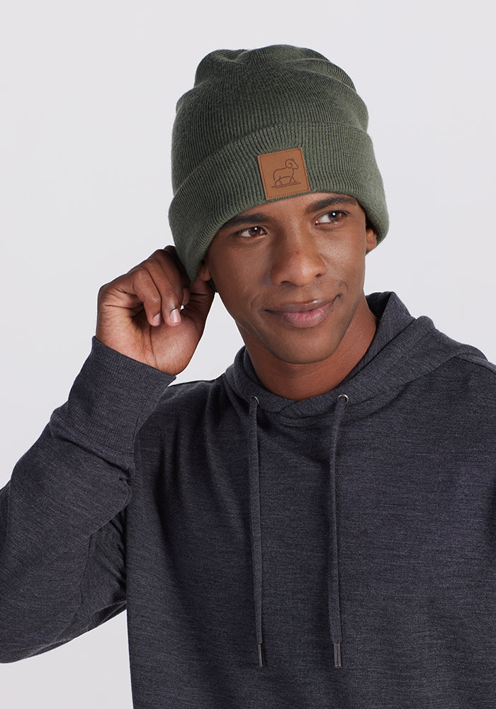 A person is posing against a plain background, wearing a dark gray hoodie and the Woolx Baylor Beanie in Spruce Heather with a small brown patch logo on the front. The person is holding one side of the beanie with a thoughtful expression.