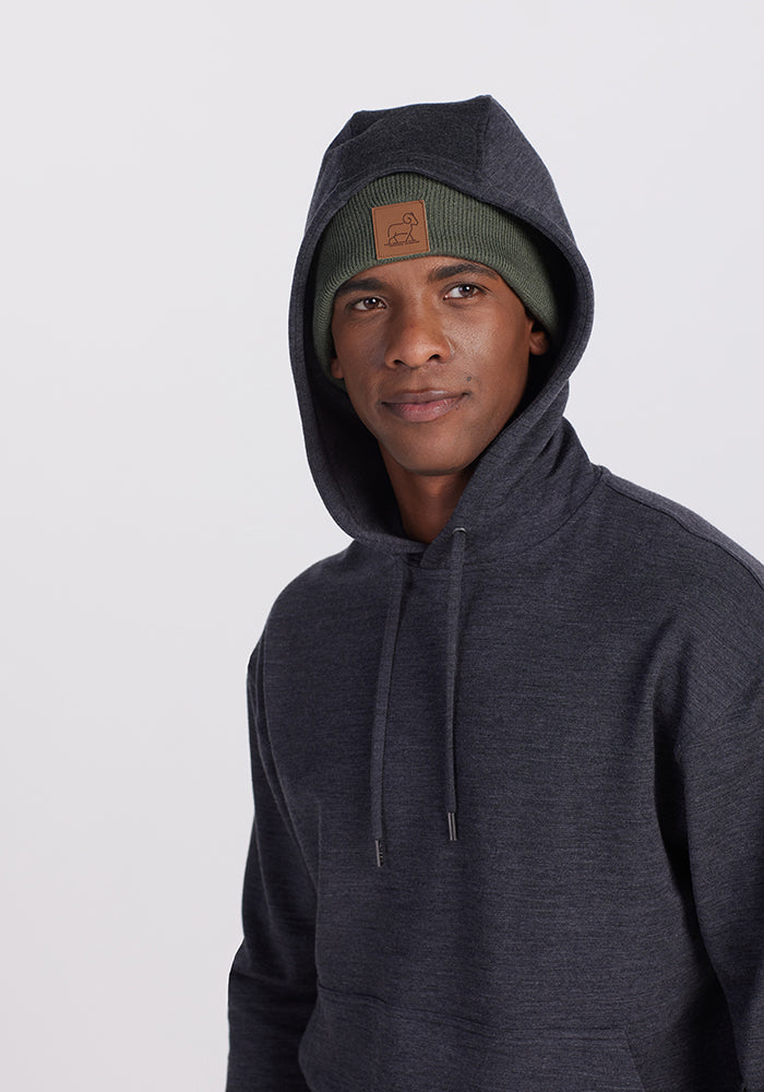 A person wearing a Woolx Chase Hoodie in Pebble Grey Melange with the hood up and a green beanie featuring a brown patch. The background is plain and light-colored.