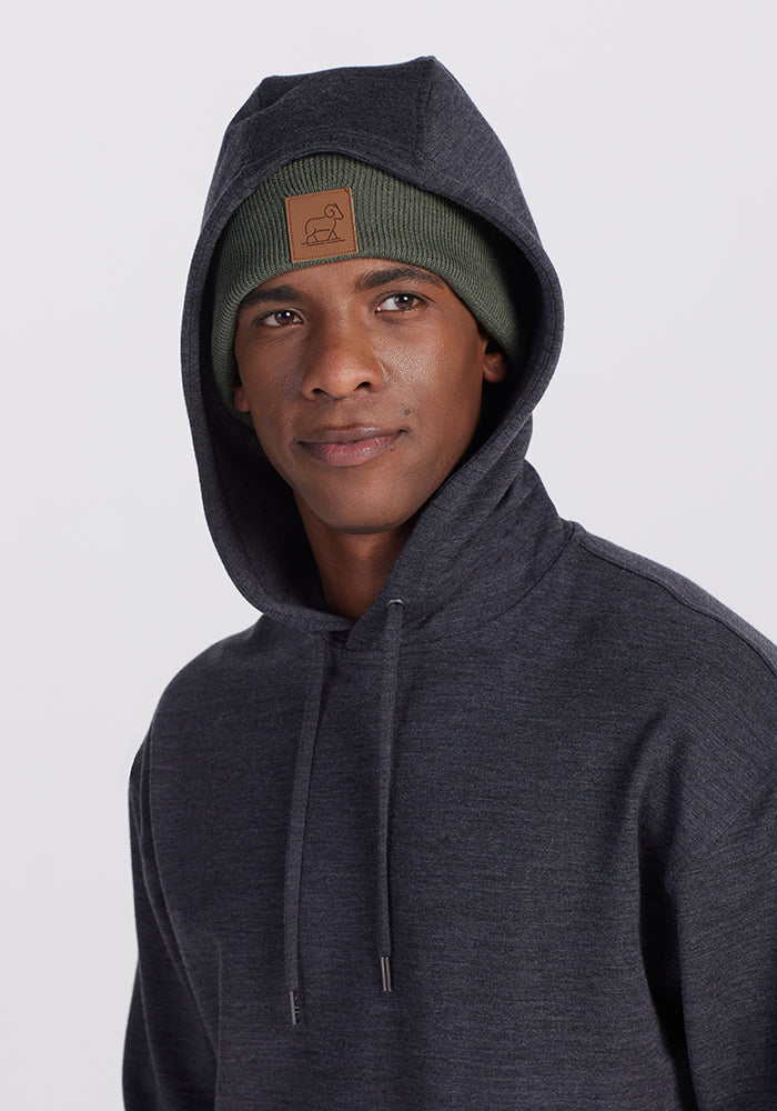 A person wearing a dark gray hoodie and a Woolx Baylor Beanie in Spruce Heather, featuring a brown patch on the front, stands in front of a plain white background, looking at the camera with a neutral expression.