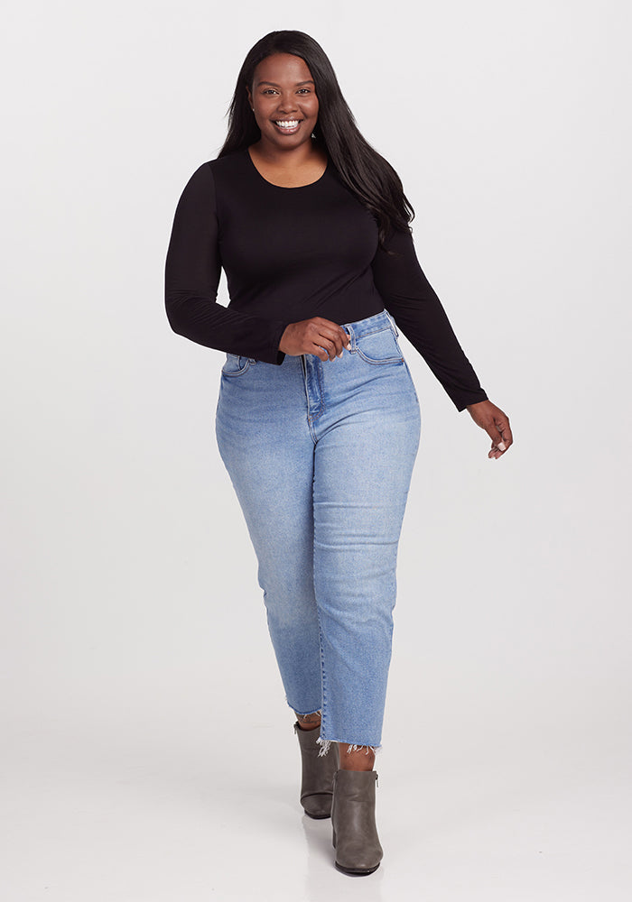 A woman is smiling while walking forward. She is wearing a long-sleeved black Sophia Bodysuit from Woolx, light blue high-waisted jeans, and gray ankle boots. The background is plain white.