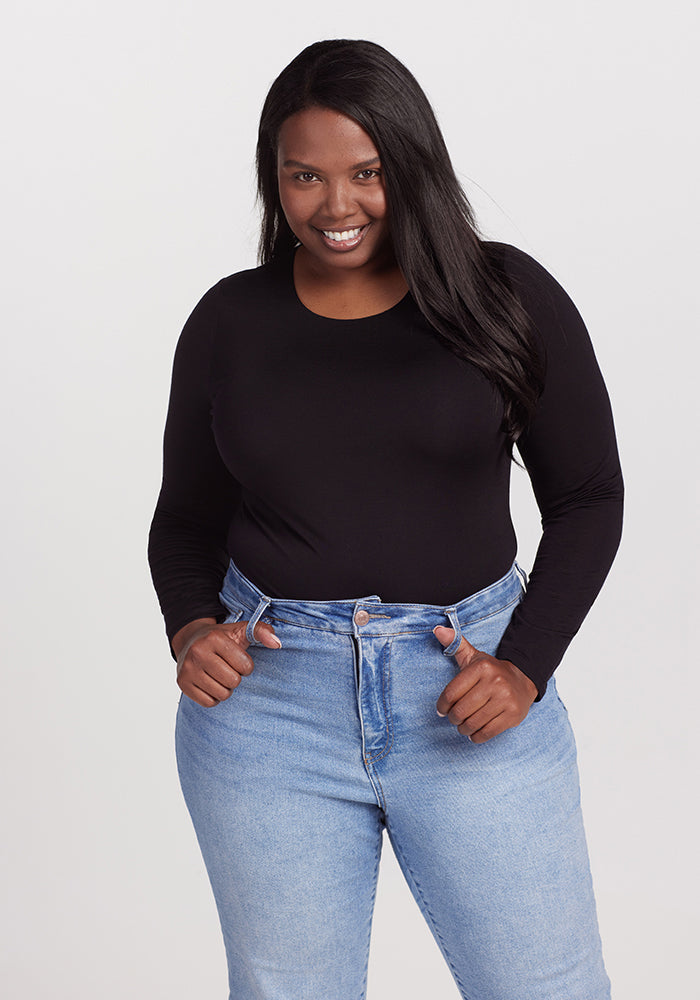 A woman with long, dark hair smiles while posing in a glamorous Woolx Sophia Bodysuit - Black and light blue jeans. She stands against a plain white background and has her hands resting on the waistband of her jeans. 