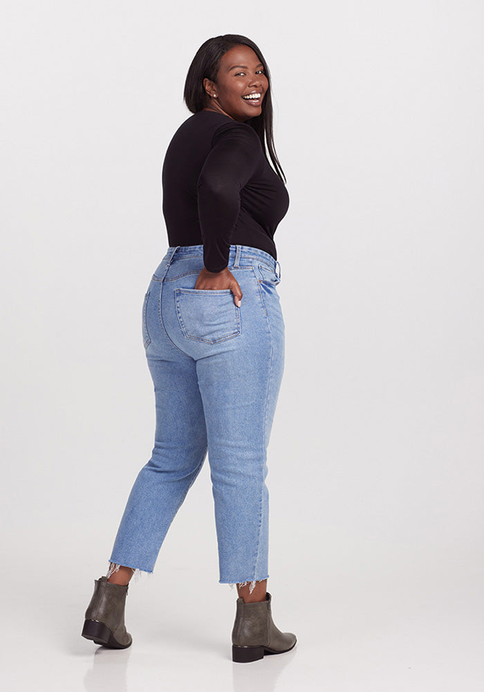 A woman with long black hair smiles while looking over her shoulder, wearing a glamorous Woolx Sophia Bodysuit - Black and light blue high-waisted jeans. She is posing with her hand in her back pocket and is also wearing gray ankle boots. The background is plain white.