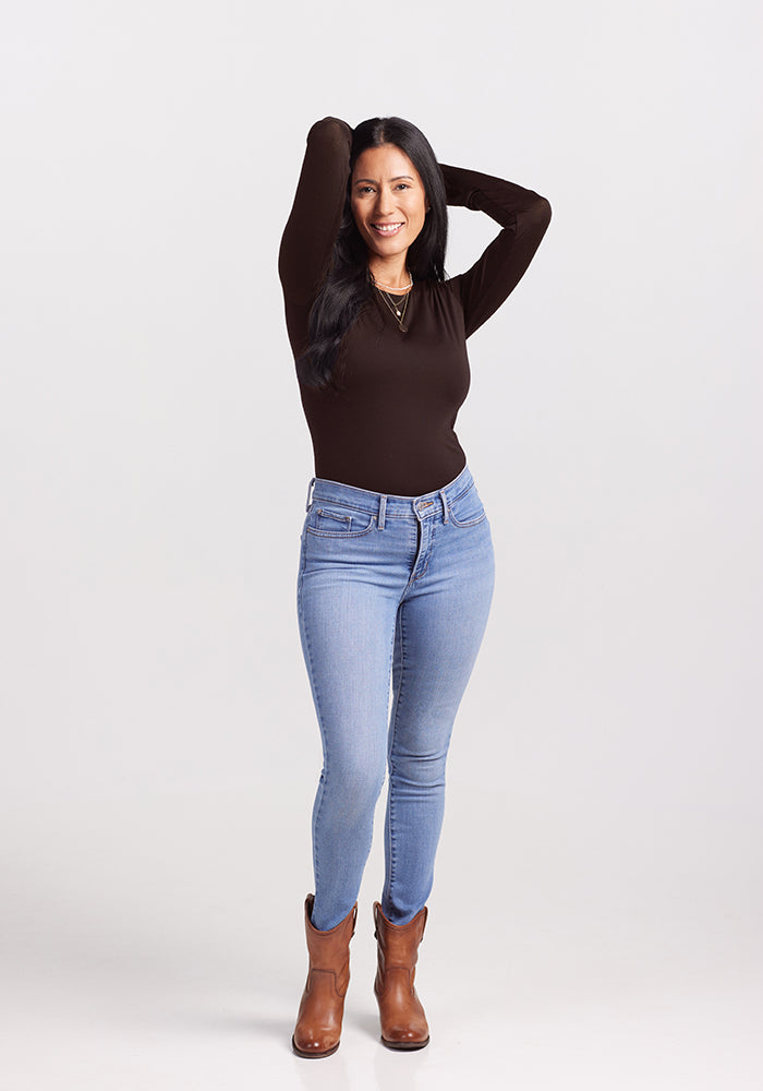 A person with long dark hair stands smiling with arms raised, showcasing a chic ensemble featuring the Woolx Sophia Bodysuit in a dark shade, light blue jeans, and brown ankle boots. The background is plain and light-colored.