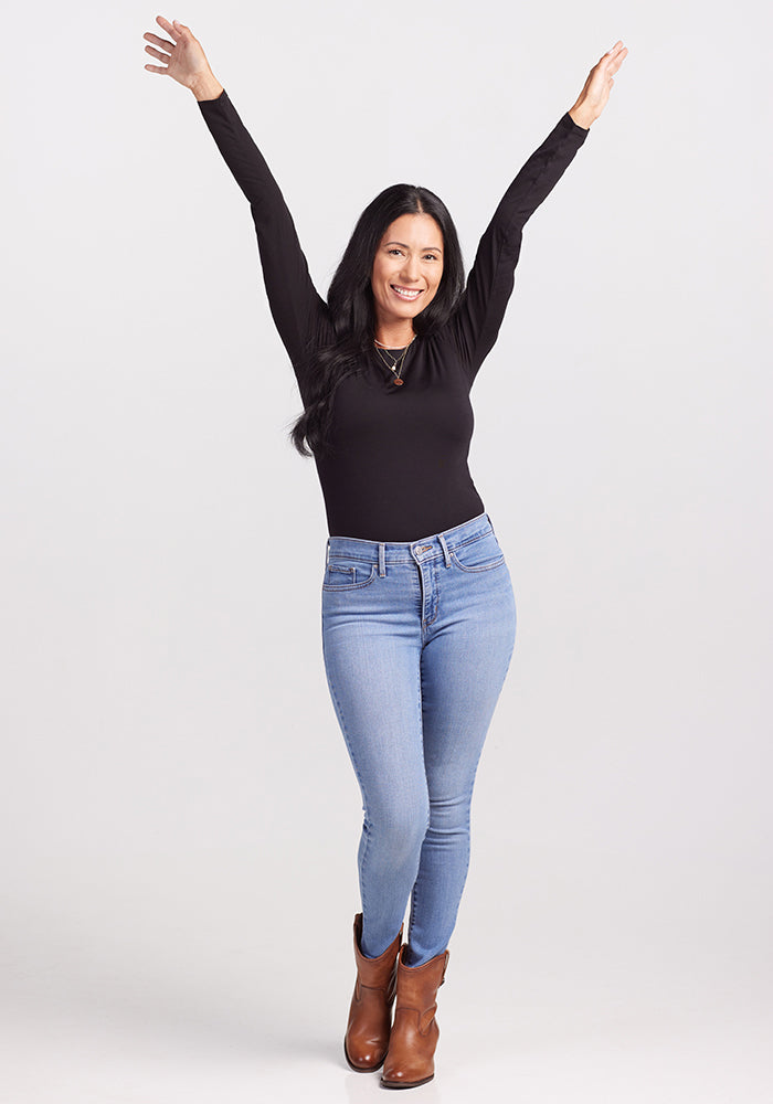 A joyful woman with long black hair stands with her arms raised, wearing a chic Sophia Bodysuit in black by Woolx, light blue jeans, and brown ankle boots. She is smiling against a plain white background.