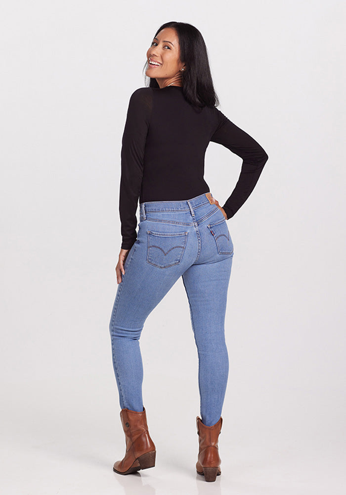 A woman standing and smiling over her shoulder, wearing a black Woolx Sophia Bodysuit, light blue skinny jeans, and brown ankle boots against a plain white background.