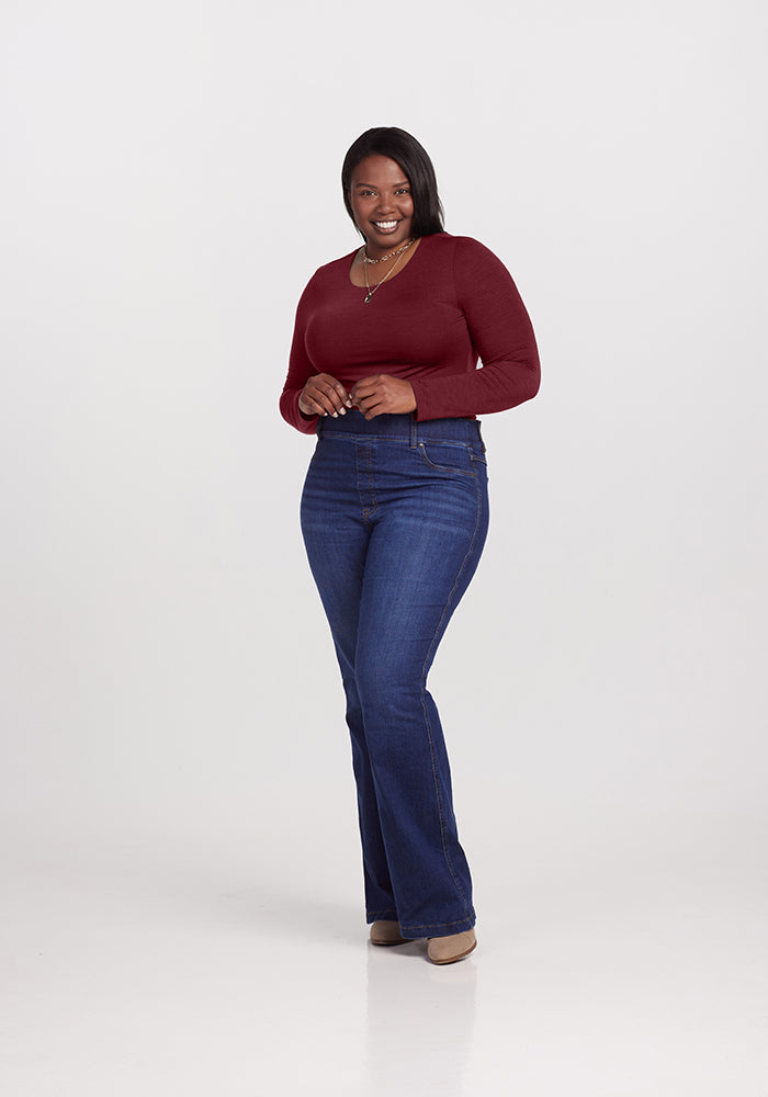 A woman stands confidently against a plain white background wearing the luxurious long-sleeved, fitted burgundy Sophia Bodysuit by Woolx, made from Australian Merino Wool. She pairs it with blue jeans that slightly flare at the bottom and accessorizes with a necklace around her neck, while her hands rest casually near her hips.