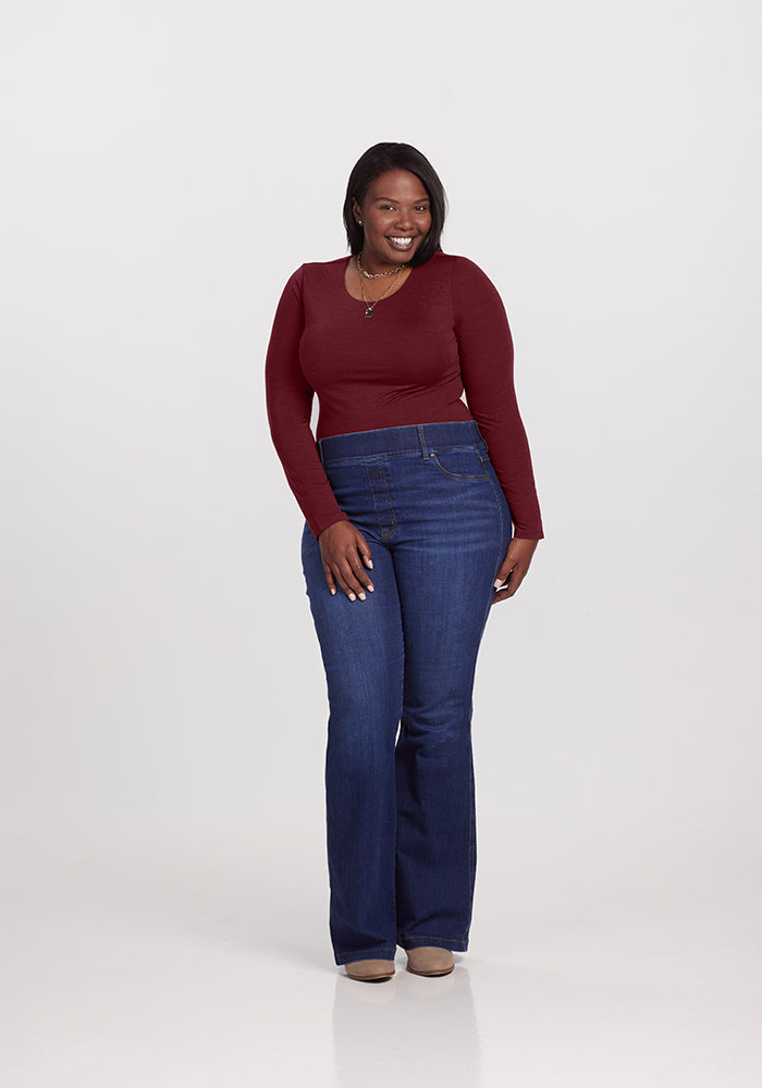 A woman stands smiling against a plain background, exuding a chic look in her long-sleeve burgundy Woolx Sophia Bodysuit, blue high-waisted jeans, and beige shoes. With dark hair cascading down, she's posing confidently with one hand on her hip and the other relaxed by her side.