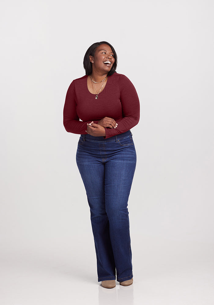 A woman with shoulder-length hair is standing against a plain white background. She is smiling and looking to her left while holding her hands together in front of her, wearing a long-sleeved maroon Woolx Sophia bodysuit, blue jeans, and beige shoes.