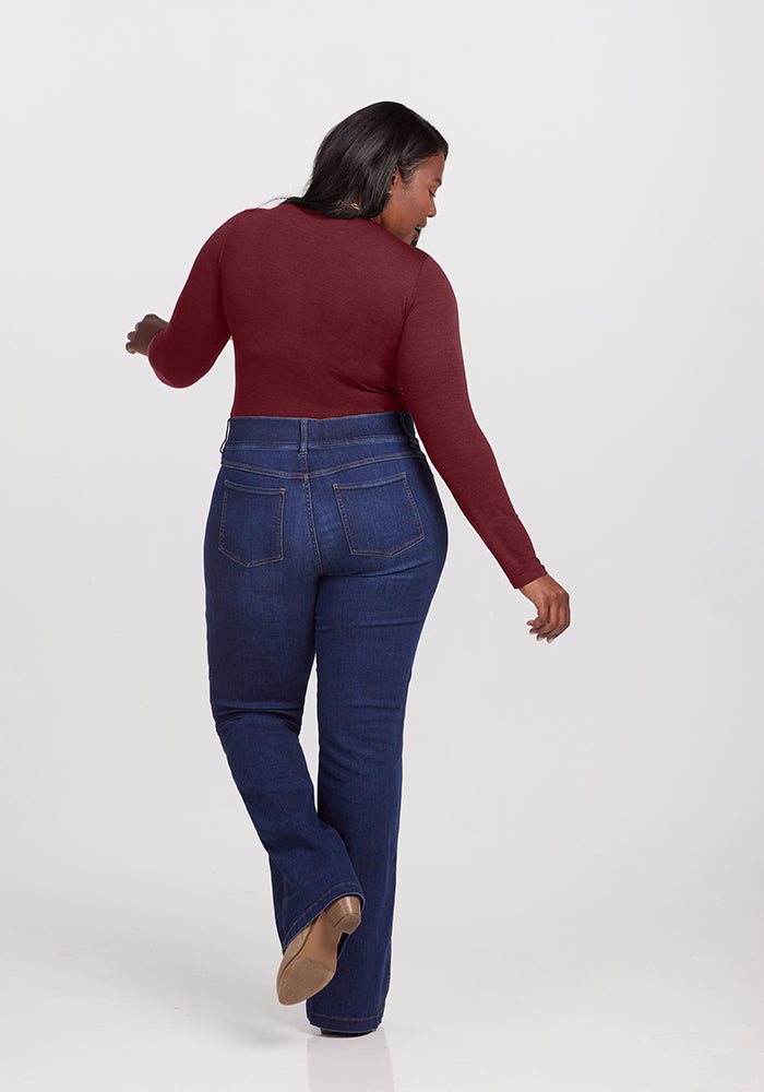 A woman is standing with her back to the camera, wearing a long-sleeve maroon Sophia Bodysuit by Woolx and dark blue jeans. She is mid-step, seemingly walking away, with straight hair cascading over her shoulders. The plain white background accentuates her chic look.