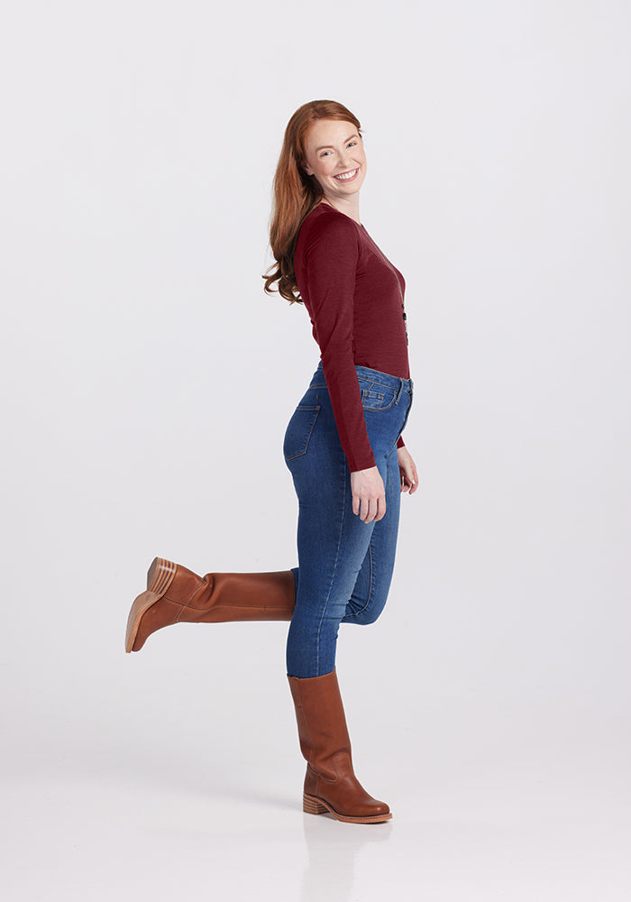 A person with long, wavy red hair is smiling and standing on one foot with the other leg bent at the knee. They are wearing a maroon Sophia bodysuit by Woolx, blue jeans, and brown boots, achieving a chic look against a plain light gray background.