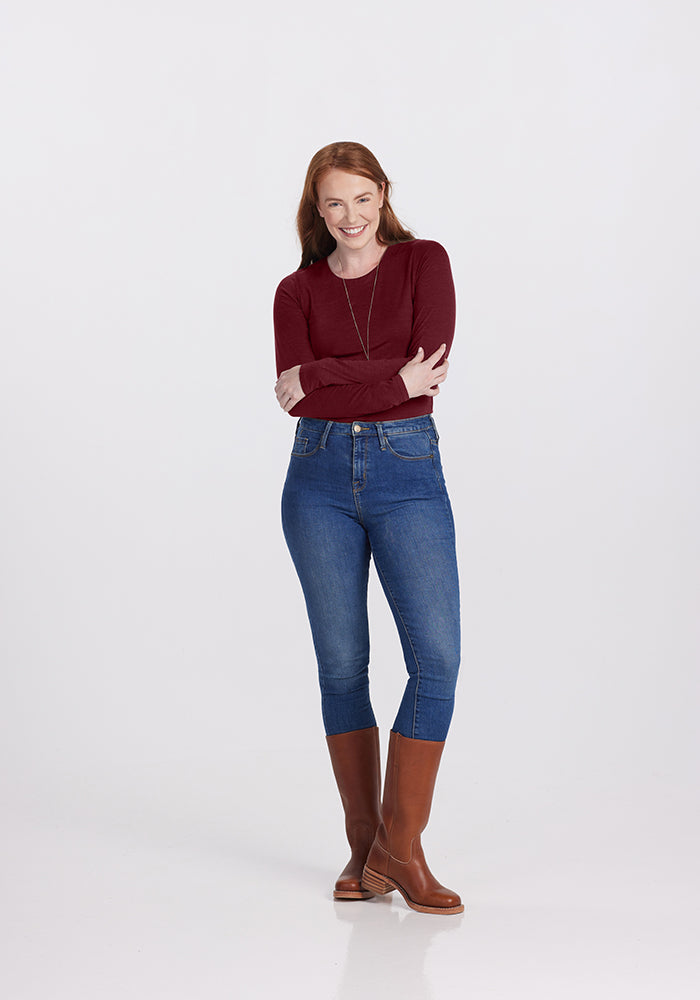 A person with long red hair stands against a plain background, smiling and looking to the side. They are dressed in a chic maroon Sophia Bodysuit made of Merino wool from Woolx, paired with blue jeans and tall brown boots. Their arms are crossed in front of them.