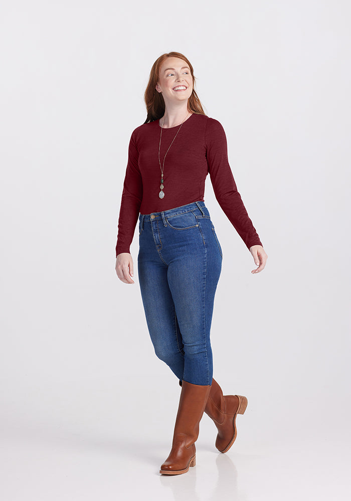 A person with long red hair walks confidently. They are wearing the chic Woolx Sophia Bodysuit, blue jeans, brown knee-high boots, and a long necklace. The background is a plain light grey.