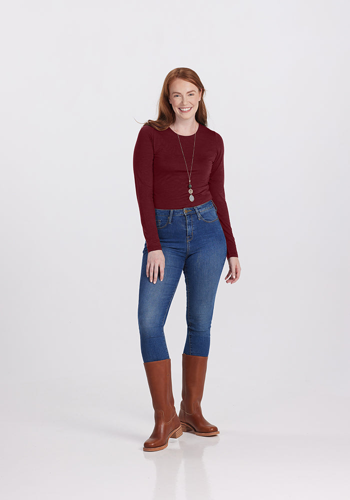 A person with long red hair is smiling and standing against a plain background. They are wearing a Woolx Sophia bodysuit paired with high-waisted blue jeans and brown knee-high boots. They have a long necklace with two circular pendants, adding to their glamorous fit.