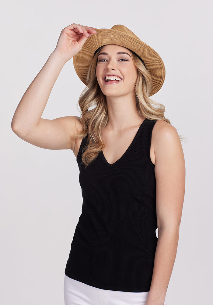 A person with long blonde hair is smiling, wearing the Woolx Vivi Ribbed Tank in black, white pants, and a straw hat. They are standing against a plain white background and touching the brim of the hat, embodying the essence of a capsule wardrobe.