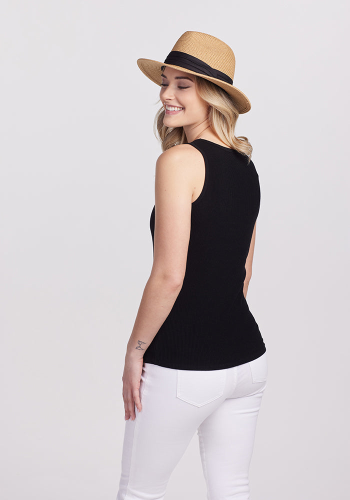 A woman wearing a straw hat and the Woolx Vivi Ribbed Tank in black smiles with her eyes closed. She is standing with her back to the camera, pairing the chic capsule wardrobe style with white pants against a plain background.