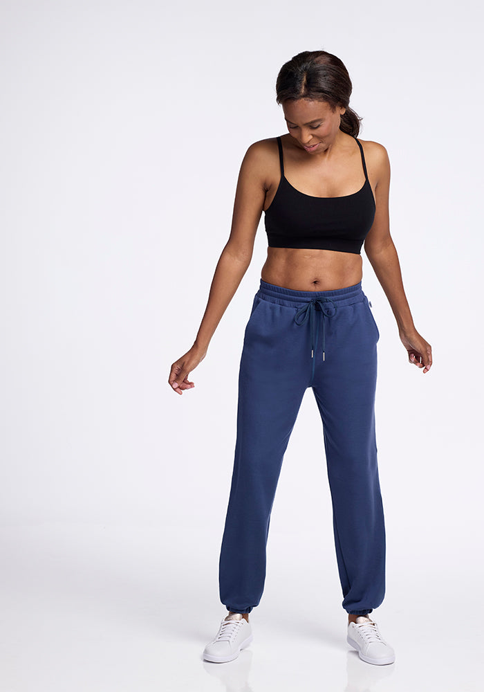 A woman in a black sports bra and blue Woolx Parker sweatpants stands against a plain white background. She looks downward with her hands by her sides, wearing white sneakers.