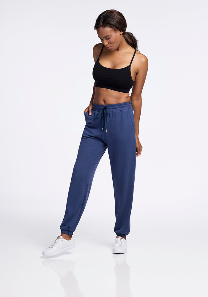 A woman is seen wearing a moisture-wicking black sports bra paired with Woolx's Parker Sweatpants in the Starry Night color, along with white sneakers. She stands against a plain white background, looking down with one hand in her pocket and the other resting on her thigh. Her hair is styled in a simple ponytail.