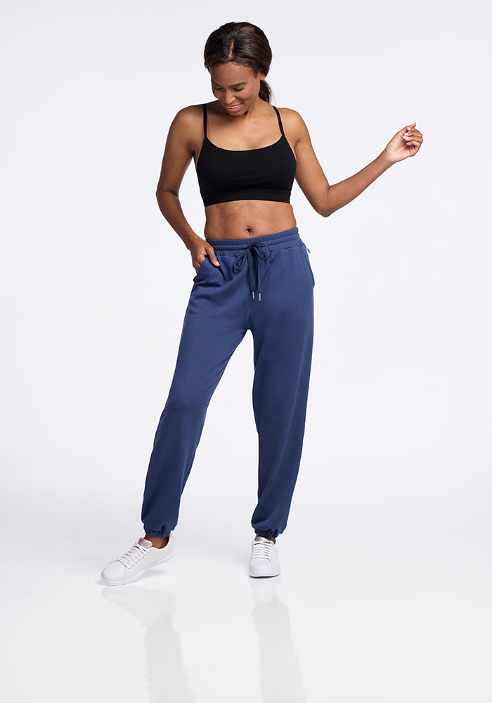 A woman stands against a plain background, wearing a black sports bra and moisture-wicking Parker Sweatpants - Starry Night from Woolx, styled in a blue jogger design and paired with white sneakers. She looks down with a slight smile, one hand on her hip and the other relaxed by her side.