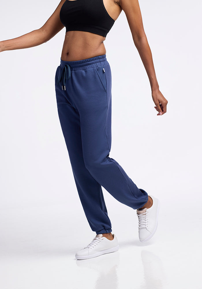 A person models Woolx's Parker Sweatpants in the Starry Night color, made from temperature-regulating Merino wool, paired with a black crop top and white sneakers. Their left arm is bent at the elbow, and the right arm is extended slightly to the side. The background is solid white, putting focus on the outfit.