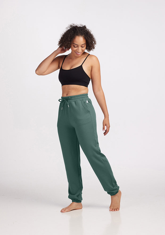 A person with curly hair wearing a black sports bra and Woolx's Parker Sweatpants in Duck Green stands barefoot on a white background, smiling and lightly touching their hair.