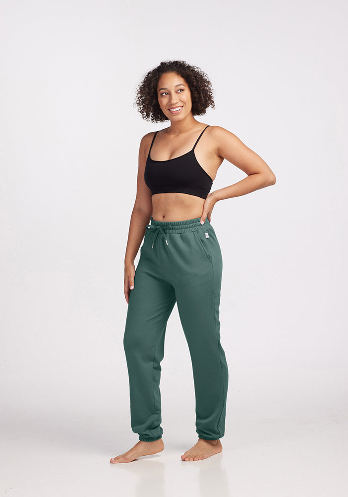 A person stands smiling against a white background, wearing a black tank top and Woolx's Parker Sweatpants in Duck Green, made from temperature-regulating Merino wool. Their left hand rests on their hip, and they are barefoot.