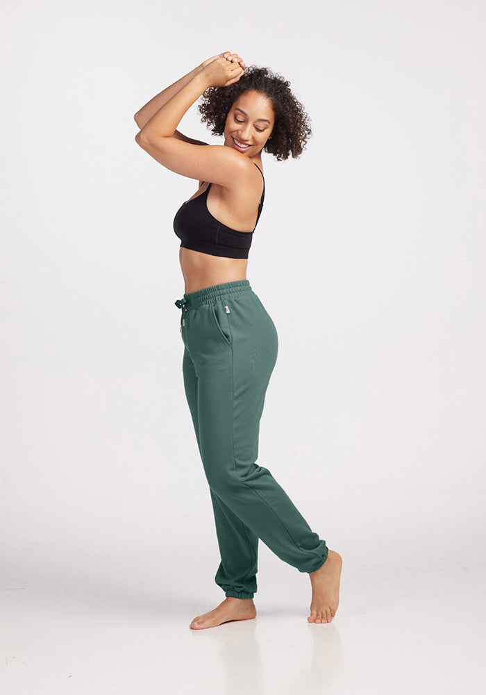 A person wearing a black sports bra and Parker Sweatpants in Duck Green from Woolx stands barefoot on a white background, smiling with arms raised and bent. The temperature-regulating outfit enhances their relaxed and joyful pose.