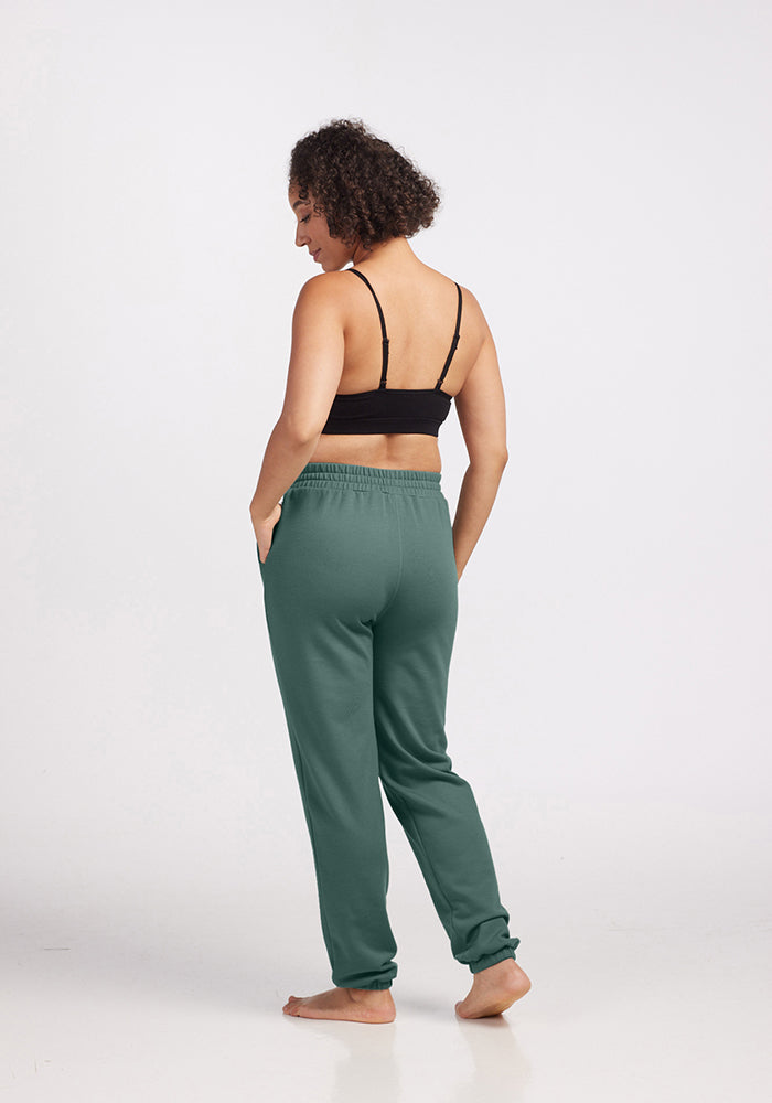 A person with curly hair stands barefoot on a white background, facing slightly away from the camera. They are wearing a black sports bra and Woolx Parker Sweatpants - Duck Green, made from temperature-regulating Merino wool.