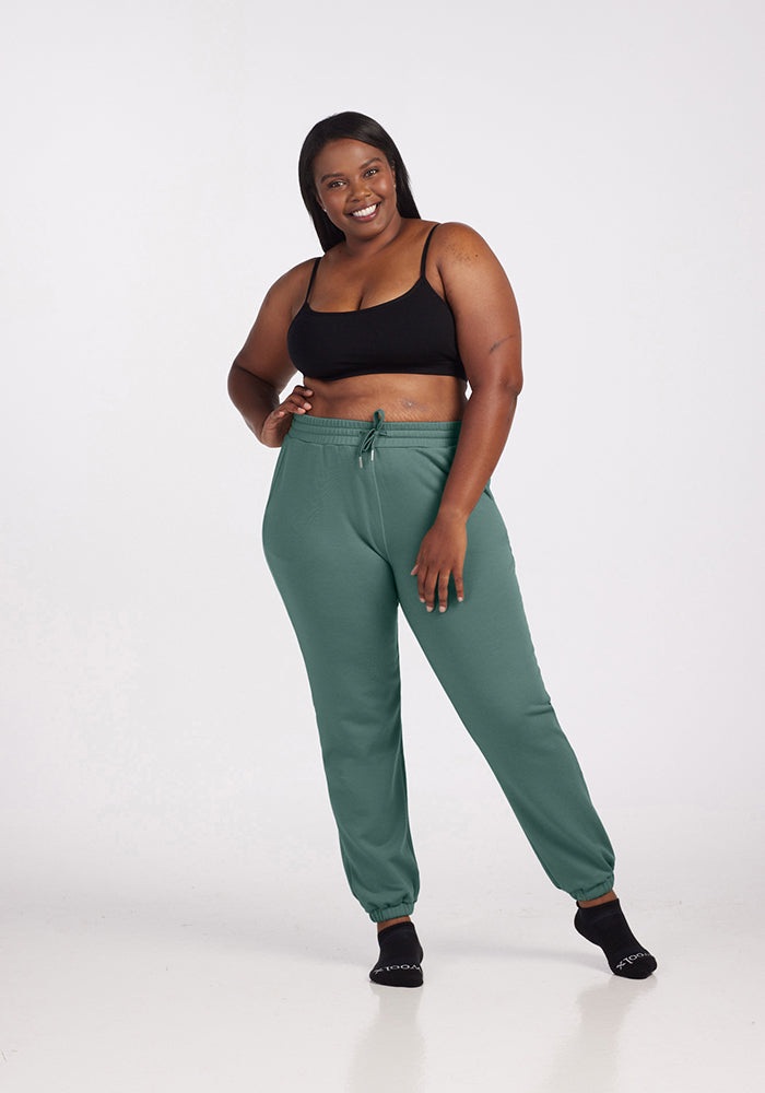 A woman smiles, standing in a relaxed pose. She is dressed in a black sports bra, Woolx Parker Sweatpants in duck green crafted from temperature-regulating Merino wool, and black socks. The background is a plain white studio setting, effectively highlighting her casual, comfortable outfit.