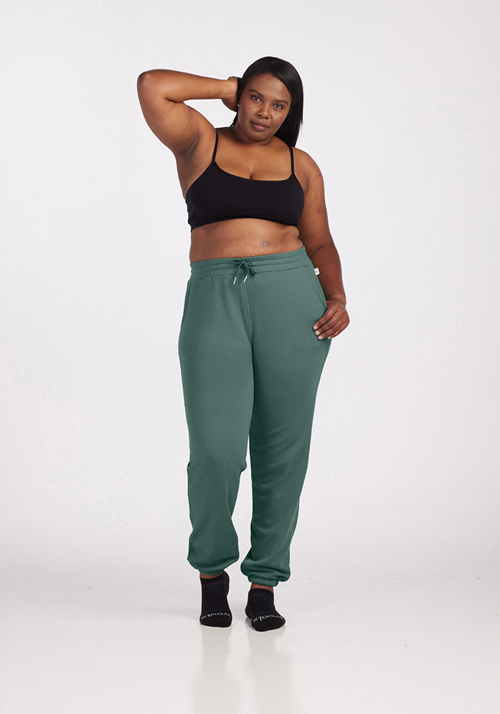 A person wearing a black sports bra and Parker Sweatpants in Duck Green from Woolx stands with one hand on their head. The temperature-regulating sweatpants pair perfectly with black socks, as they pose barefoot against a plain white background.