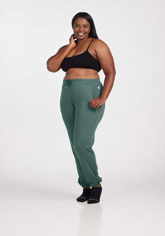 A person is smiling while posing in Woolx's Parker Sweatpants in Duck Green and a black crop top. They are standing against a plain white background, resting one hand on their cheek, exuding comfort and confidence in their odor-free outfit.