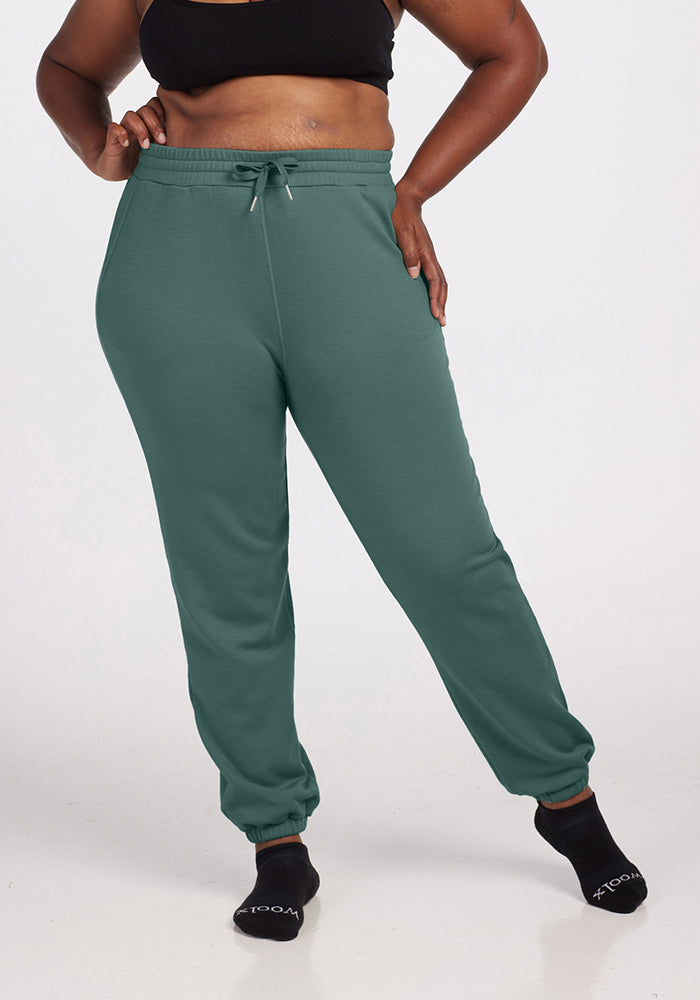 A person wearing a black sports bra, Woolx's Parker Sweatpants in Duck Green with a drawstring waist, and black socks stands against a plain background. These temperature-regulating sweatpants ensure an odor-free experience all day long.